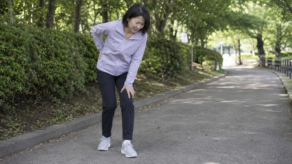 脊柱管狭窄症で歩行中に足の痛みとしびれを感じ、立ち止まって腰と膝に手を当てる女性(間欠性跛行のイメージ)