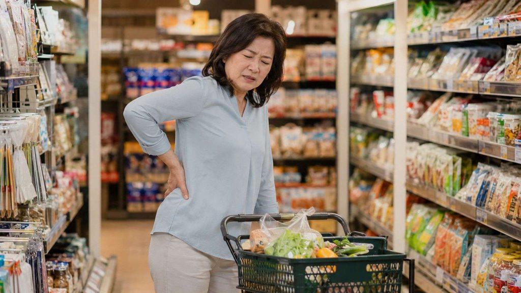 買い物中に前かがみの姿勢で腰に手を当て、痛みをこらえる女性 脊柱管狭窄症の症状